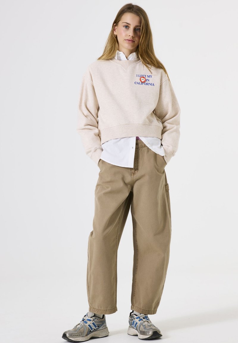 Lichtbeige cropped sweatshirt met print, gelaagd over een witte shirt. Ruimvallende khaki broek en grijze sneakers met blauwe accenten.