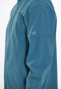 Tealfärgad jacka med en slät, matt yta. Den har hög krage, långa ärmar och en subtil broderad logotyp på ärmen.