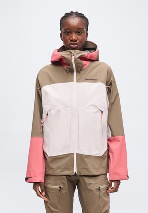 Personne portant une veste d'extérieur color block beige, blanche et rose avec capuche et fermeture éclair à l'avant, debout devant un fond clair uni.