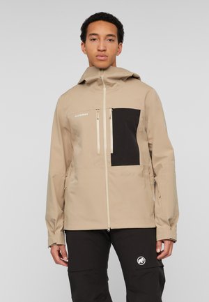 Young person wearing a beige jacket with black chest pocket and black pants with a white logo on the thigh, facing forward against a gray background.