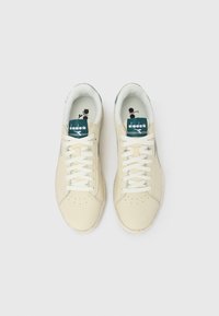 Ζευγάρι μπεζ αθλητικά παπούτσια Diadora με λευκά κορδόνια και ετικέτες στη γλώσσα σε σκούρο πράσινο, από πάνω σε λευκό φόντο.