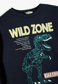 Sudadera azul marino con un diseño geométrico de T. rex en teal y el texto "WILD ZONE". Tela texturada con cuello acanalado. Acentuada con "ROAR KING."