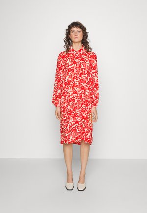 Vestido com estampado floral vermelho e branco, com mangas longas, colarinho abotoado, comprimento intermédio, corte relaxado e fendas laterais. Usado com sapatos de salto bloco bege.