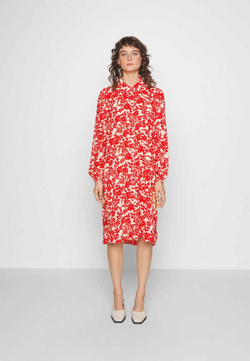 Vestido com estampado floral vermelho e branco, com mangas longas, colarinho abotoado, comprimento intermédio, corte relaxado e fendas laterais. Usado com sapatos de salto bloco bege.