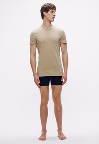 Μπεζ βαμβακερό t-shirt με στρογγυλή λαιμόκοψη, σε συνδυασμό με σκούρο μπλε σορτς. Απλός σχεδιασμός, εφαρμοστή γραμμή, λεία υφή και κοντά μανίκια.