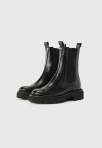 Des bottes Chelsea en cuir noir présentent des panneaux latéraux en élastique, une semelle en caoutchouc plate avec une texture antidérapante et des languettes à l'arrière pour un enfilage facile.