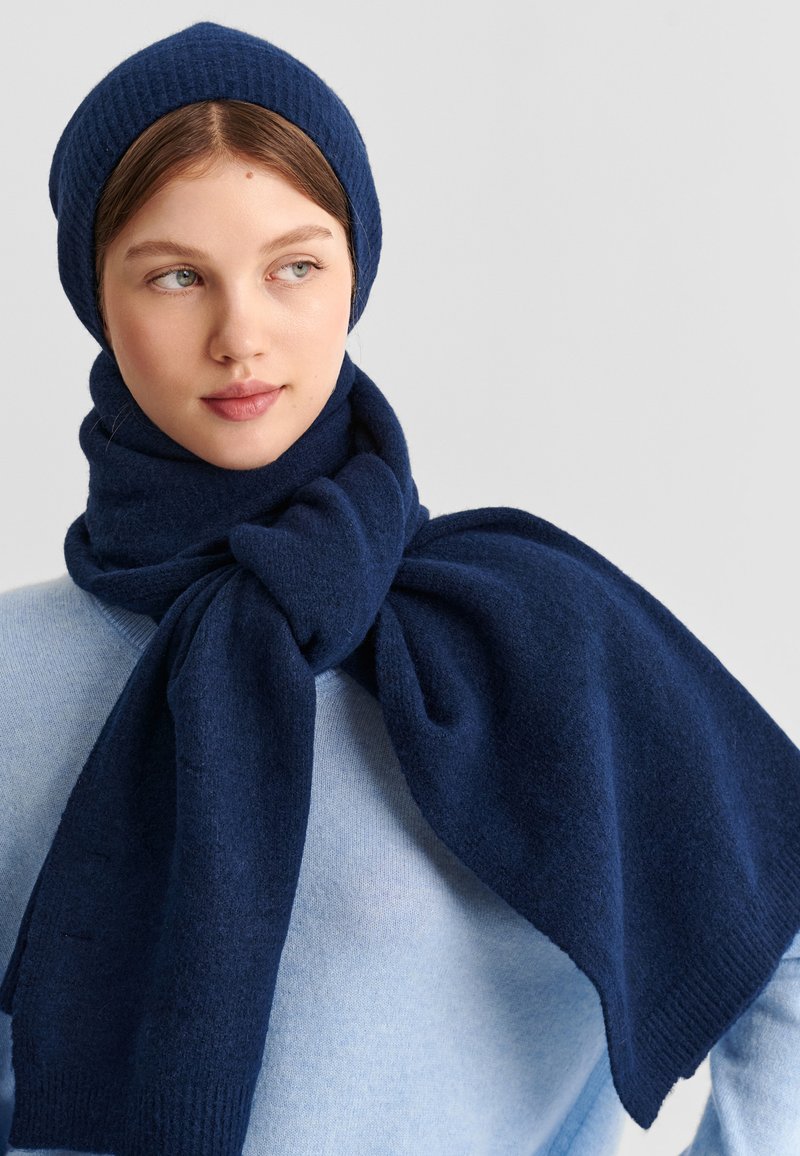 Jeune femme à la peau claire et aux cheveux châtain clair portant un bonnet en maille bleu marine et une écharpe assortie sur un pull bleu clair, regardant sur le côté.