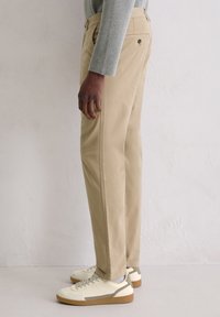 Beige pants made of cotton with a straight-leg design, side pockets, and back buttoned pockets, paired with white and gray sneakers.