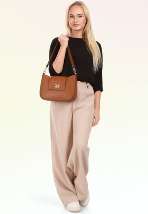 Blondine vrouw die een zwarte top en beige wijde broek draagt, houdt een bruine leren handtas met een gouden gesp op haar schouder.