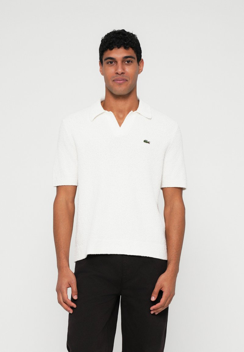 Polo-shirt blanc à manches courtes avec une finition texturée et un col à encolure en V. Présente un petit logo vert sur la poitrine.