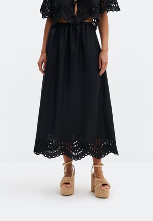 Žena v čiernej midi sukni s kvetinovým vyrezávaným lemom a béžovými pletenými platformovými sandálmi stojaca na jednobielom pozadí.