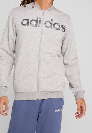 Felpa grigia con zip, colletto alto e polsini a coste. Presenta un grande logo Adidas nero sul petto in un motivo camouflage.