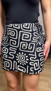 Femme portant une jupe portefeuille noire ornée de motifs géométriques blancs et de motifs solaires, associée à un haut noir ajusté, debout sur un sol en bois clair.