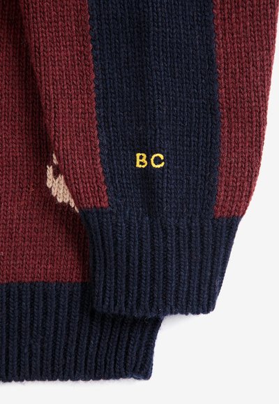 Gros plan sur des manches de pull tricoté avec des rayures bleu marine et bordeaux et des lettres jaunes brodées "BC" sur la partie bleu marine.