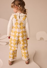 Salopette à carreaux jaunes et blancs avec bretelles, associée à un haut ivoire à manches longues avec un col à volants et un détail de boutons.