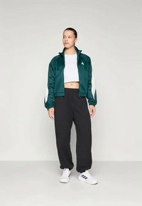 Πράσινο σατέν μπουφάν με λευκές λεπτομέρειες, συνδυασμένο με μαύρο παντελόνι φαρδιάς γραμμής και λευκά αθλητικά παπούτσια. Το μοντέλο στέκεται όρθιο μπροστά σε ουδέτερο φόντο.