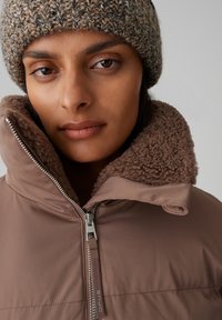 Veste matelassée brune avec un col texturé et duveteux. Dotée d'une fermeture éclair argentée et d'un tissu doux à la finition lisse. Complétée par un bonnet en tricot.