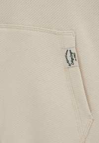 Világos bézs szövet texturált kötött mintával. Zseb és egy "College League" feliratos címke található rajta, az 1962-es évszámmal.