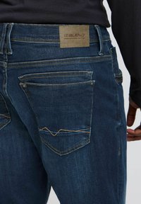 Denim jeans i mørk blå med en jevn tekstur, med et skinnlapp merket "BLEND," og kontrasterende oransje søm på baklommene.