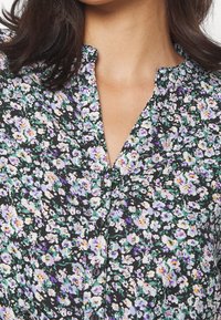Femme portant un chemisier noir à motifs floraux violets, blancs et verts, avec un col rond et des boutons noirs sur le devant.