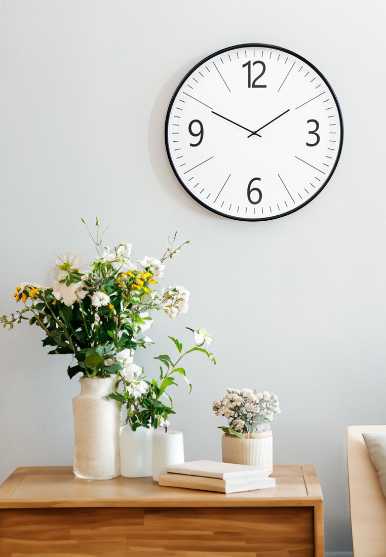 Schwarze Wanduhr mit weißem Zifferblatt und fetten schwarzen Ziffern, hängt über einem Holztisch mit Blumenvasen und gestapelten Büchern.