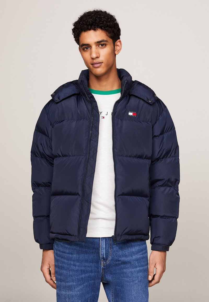 Giacca puffer blu navy con zip frontale, cappuccio removibile e logo sul petto. Presenta una texture trapuntata e polsini elasticizzati.