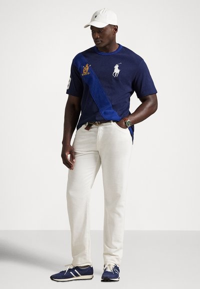 Πόλο πουκάμισο σε μπλε και ναυτικό μπλε με κεντημένο λογότυπο, συνδυασμένο με λευκό τζιν και μπλε αθλητικά παπούτσια, ολοκληρωμένο με λευκό καπέλο.