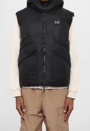 Osoba ubrana w czarną pikowaną kamizelkę bez rękawów z przednimi kieszeniami, na kremowej bluzie oraz beżowe spodnie typu cargo.