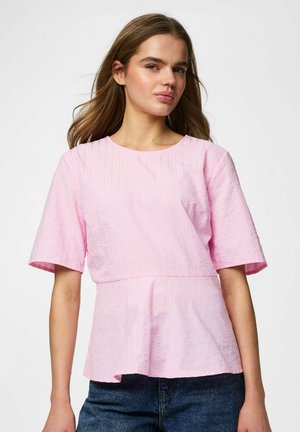 Ung kvinde med langt hår iført en lyserød kortærmet bluse med subtile lodrette mønstre og blå jeans foran en ensfarvet baggrund.