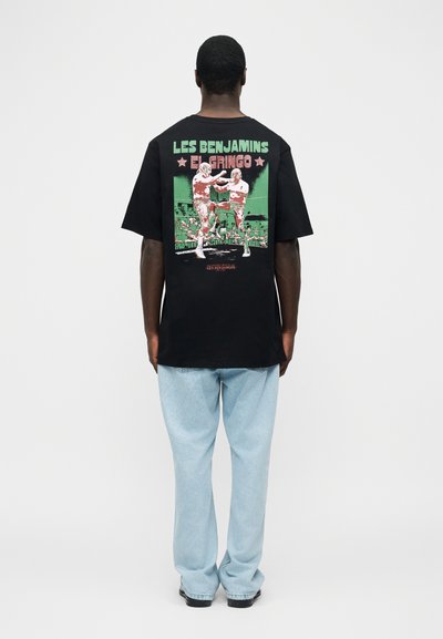 Άνδρας με γαλάζια τζιν και μαύρο μπλουζάκι με γραφικό δύο παλαιστών που αγωνίζονται και κείμενο "LES BENJAMINS EL GRINGO" στην πλάτη, όρθιος με την πλάτη στραμμένη προς το μέρος μας.