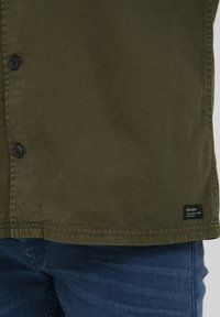Green button-up shirt made of cotton fabric, featuring black buttons, a straight hem, and a small brand tag at the bottom right.