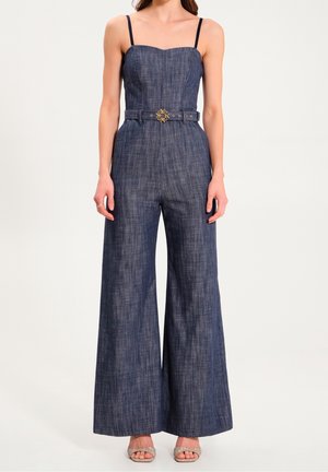 Salopette en denim foncé avec des bretelles fines, un corsage ajusté, un pantalon à jambes larges et une taille ceinturée avec un détail métallique décoratif.