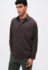 Pruunist ruuduline nööpidega särk krae, pikkade varrukatega ja pooke-taskuga; kombineeritud oliivroheliste pükstega.