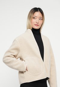 Γυναίκα που φοράει μπεζ φλις τζάκετ πάνω από μαύρο πουλόβερ με γιακά, με το ένα χέρι της στην τσέπη του τζάκετ, κοιτάζοντας πλάγια σε έναν απλό φόντο.