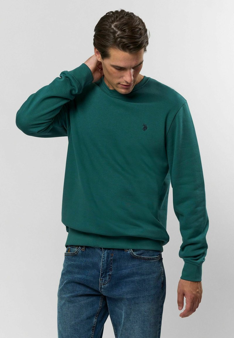 Zöld crew nyakú pulóver sima anyagból, hosszú ujjakkal és egy kicsi logóval a mellkason. Kék farmerral párosítva.