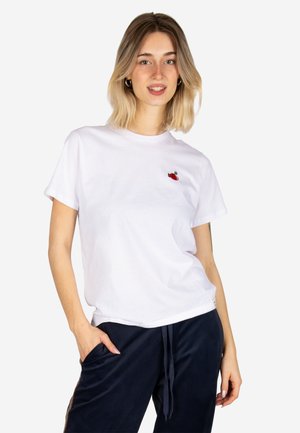 Junge Frau mit schulterlangem blondem Haar, die ein weißes T-Shirt mit kleinem roten Herzmotiv und dunkle Hose trägt, lächelt mit einer Hand in der Tasche.