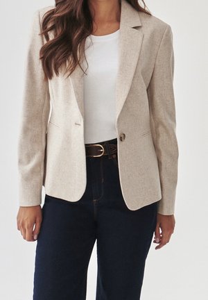 Vrouw draagt een beige blazer over een wit shirt met donkere spijkerbroek en zwarte riem, staand tegen een effen lichte achtergrond.