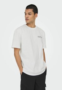 T-shirt en coton blanc à manches courtes et col ras du cou. Présente un petit logo noir sur la poitrine gauche. Porté avec un short cargo noir.