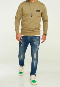 Uomo che indossa un felpa verde oliva, jeans blu scoloriti, sneakers verdi e bianche, e una collana con un dog tag nero.