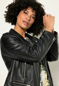 Veste en cuir noir avec des zips en argent, présentant un design structuré, une texture légèrement brillante et des accents nervurés le long des épaules.
