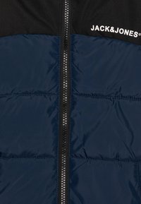 Črna in mornarsko modra puhovka z zadrgo spredaj, horizontalnim kvadratnim vzorcem in belim logotipom "JACK & JONES" v zgornjem delu.