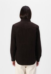 Camicia di velluto a coste marrone con colletto, texture a righe verticali e polsini con bottoni, caratterizzata da un orlo curvo e un dettaglio sullo yoke sul retro.