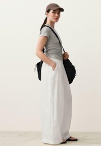Femme portant une casquette de baseball taupe, un t-shirt gris, un pantalon blanc à jambes larges, un sac à bandoulière noir et des sandales noires, debout de profil sur un fond uni.