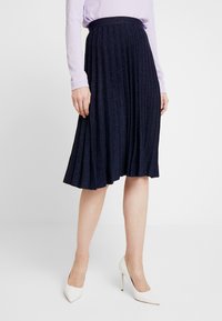 Marinblå plisserad kjol med en glittrig textur, utrustad med ett enhetligt midjeband, i kombination med vita stilettklackar och en ljuslila topp.