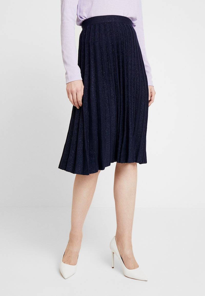 Marinblå plisserad kjol med en glittrig textur, utrustad med ett enhetligt midjeband, i kombination med vita stilettklackar och en ljuslila topp.