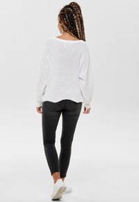 Weißer Strickpullover mit lockerem Schnitt, weiten Ärmeln und abgerundetem Saum, kombiniert mit dunklen Jeans und weißen Sportschuhen.