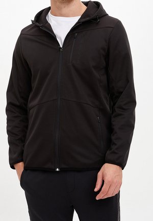 Mann trägt eine schwarze Kapuzenjacke mit Reißverschluss über einem weißen Hemd, die Hände in den Jackentaschen vor einfarbigem Hintergrund.