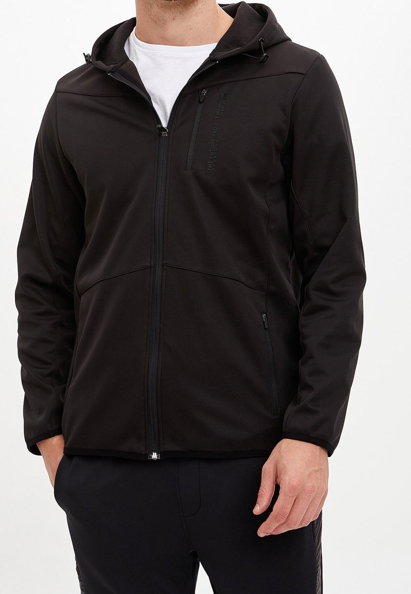 Homme portant une veste à capuche noire zippée sur une chemise blanche, les mains dans les poches de la veste, sur un fond uni.