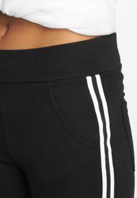 Pantalons de sport noirs avec une large ceinture élastique, une poche latérale et deux bandes blanches verticales le long des coutures extérieures.