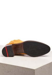 Žluté semišové boty s plochou, texturovanou černou gumovou podrážkou. Výrazný červený akcent na patě a hnědé kožené podšívce. Disponují bezešvým designem.